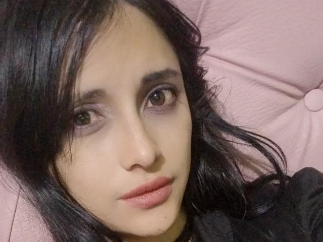 MidoriChang's face, showing dark wavy hair, purple eyeshadow, brown eyes, and pink lips, lying on a pink pillow. She's wearing a black top.