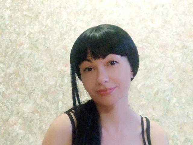 ReifeVennity-hot hat schwarzes Haar, eine Pony und einen seitlichen Pferdeschwanz, trägt ein schwarzes Top mit Trägern, hat ein leichtes Lächeln und helle Haut, auf einem einfachen Foto mit weichem Licht..