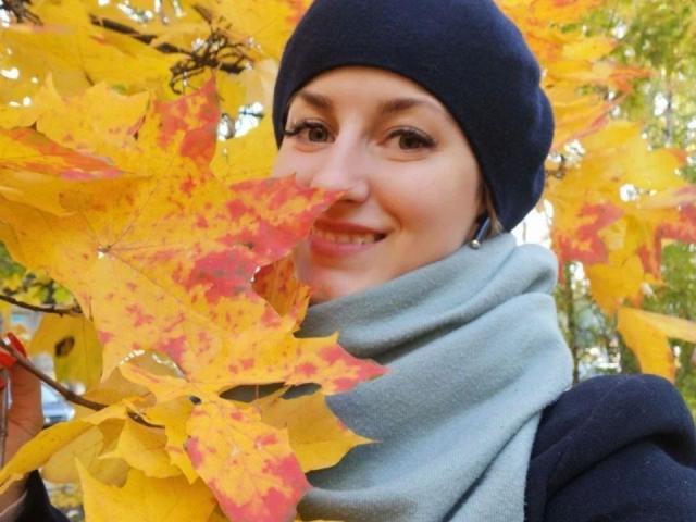 ChristineLove-hot with yellow autumn leaves in her face, wearing a dark blue beanie and light gray scarf, smiling with brown eyes. Her ass is covered by her scarf.