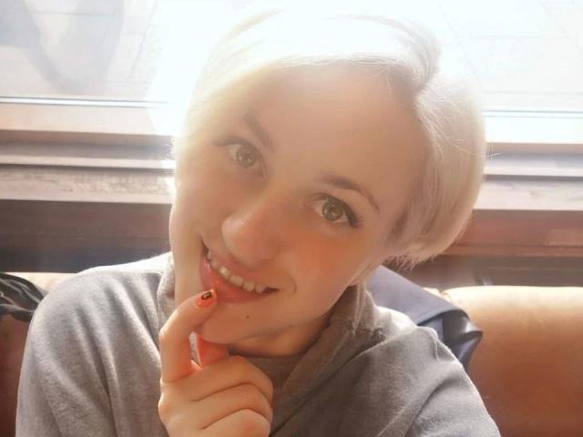 ChristineLove-hot with short blonde hair, smiling, touching her finger to her lips with a cute, small red nail, wearing a gray shirt, in a bright, sunny setting.
