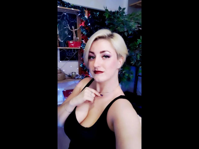 ChristineLove-hot with short blonde hair, wearing a black tank top, showing off her big tits, red lipstick, and a confident smile, touching her neck, in a cozy room.
