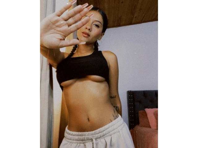 ThaliaAres with tan skin, small tits, and tattoos, wearing a black sports bra and gray sweatpants, covering her face with her hand, in a wooden ceiling room.