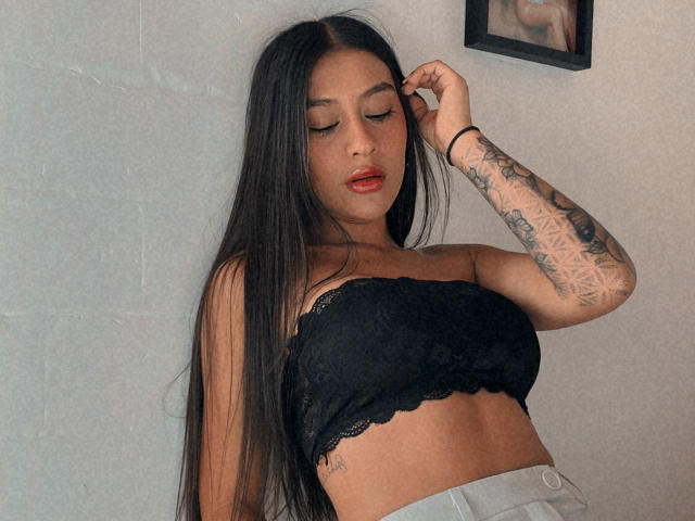 ThaliaAres with long black hair, wearing a black lace strapless top, showing tattoos on her right arm, and light gray pants, eyes closed, hand in hair, red lips.