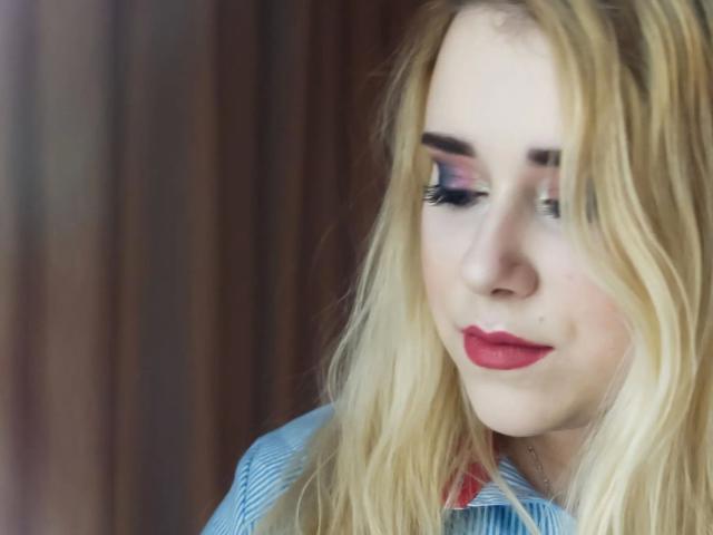 HaileyBLONDE-hot with blonde hair, red lipstick, and blue eyes, wearing a striped shirt, looking down with a slight smile. Boobs not visible. Soft, blurred background.