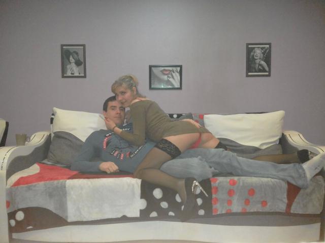 Bobandchris33-hot with blonde hair, green dress, and black stockings, leans over bobandchris33-hot in gray sweater, red beads, on patterned couch, showing bare ass.