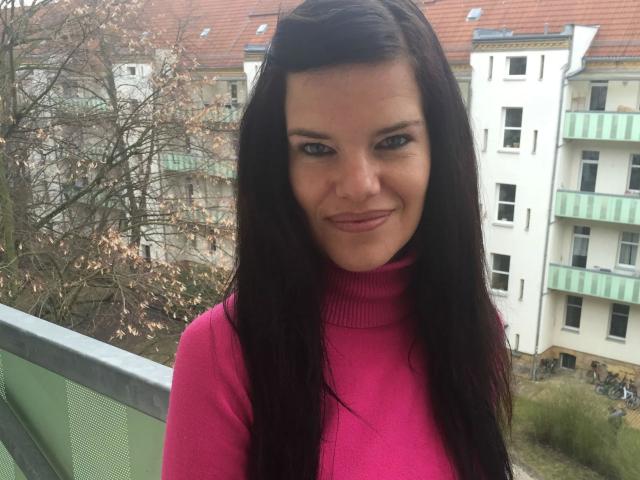 Jenny-Sun with dark brown hair, pink turtleneck, and a slight smile, showing her tits in the photo, standing outside with a green railing and white building behind.