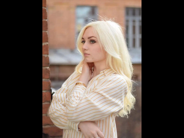 EvaTesla-hot with long blonde hair, wearing a striped shirt, crosses arms, touches chin, looking serious, gold bracelet, standing against a brick wall.