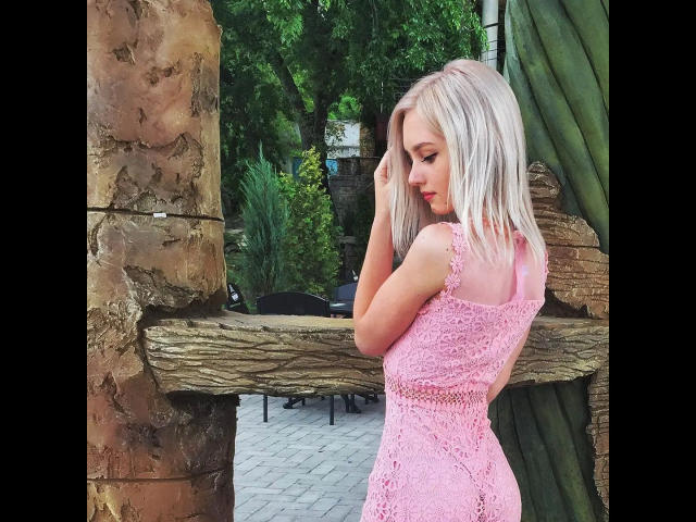 EvaTesla-hot with pale pink lace dress, showing her ass, standing in front of wooden rock structure, green leafy background, blonde hair, looking down, hand in hair.