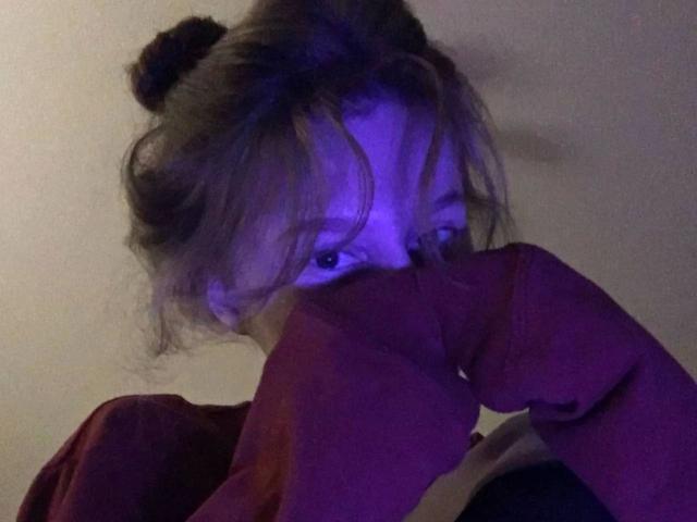 Enigma-hot with messy brown hair, blue eyes, and a red long-sleeve shirt, biting her forearm, looking up with a slightly curious expression.