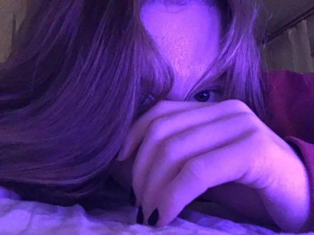 Enigma-hot with purple-tinted hair, eyes peeking through fingers, black nail polish, lying on bed, partially covered face, soft lighting, relaxed expression.