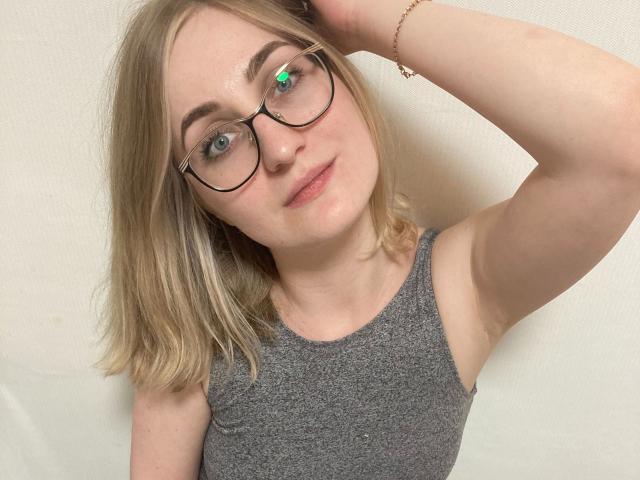 Kristinakokietka-hot with blonde hair, glasses, and a gray sleeveless top, raising her right arm, showing her armpit, and wearing a gold bracelet, has green eyes and a slight smile.