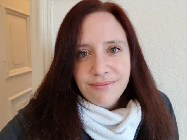 JessicaGold-hot has pale skin, green eyes, and brown hair. She's wearing a white scarf and black jacket, smiling softly with pink lips. Her tits are not visible.