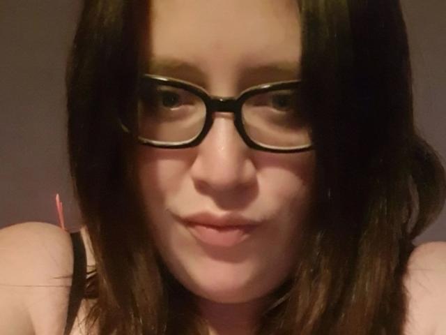 JessicaGold-hot with dark brown hair and black glasses, showing a slight smile, with her pink nipples barely visible in a black strap top.