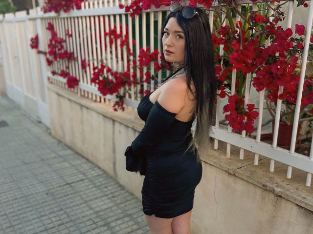Claudiliciousxx-hot with long dark hair, black off-shoulder dress, and sunglasses on head, standing by white fence with red flowers, looking over shoulder, showing her ass and tits.
