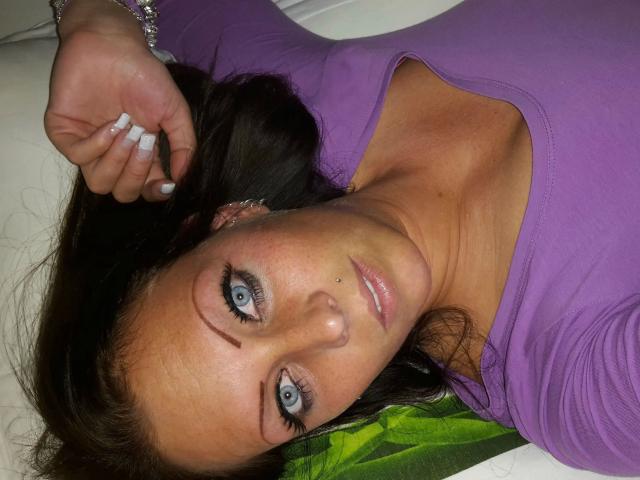 HotNaisha-sex with blue eyes, dark brown hair, purple top, nose piercing, and white nails, lying down, looking up, wearing a purple bracelet, on a white surface.