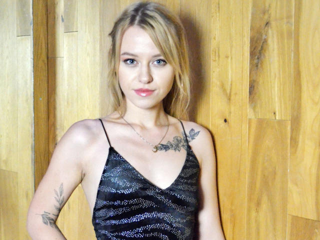 Cindy-Lust with blonde hair, blue eyes, and pink lipstick, wearing a sparkly black dress, showing tattoos on her chest and arm, standing against wooden panels.