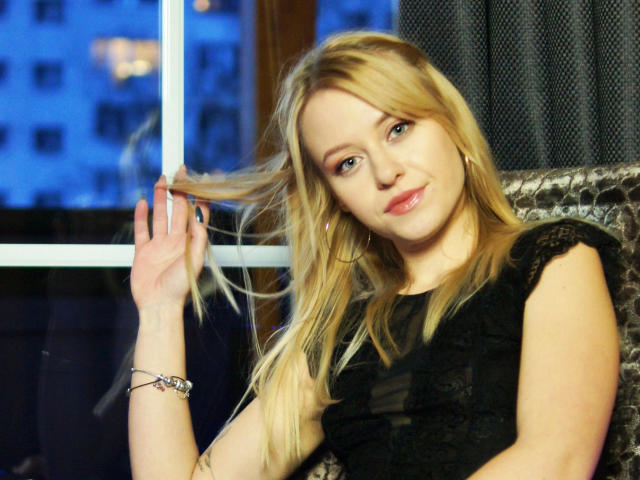 Cindy-Lust with blonde hair, holding a lock, wearing a black top, small tits, light skin, pink lips, silver bracelet, sitting by a window, blue city lights outside.