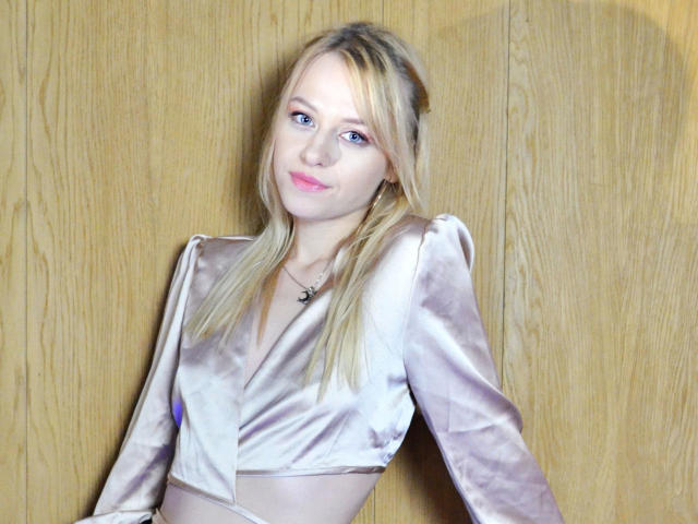 Cindy-Lust has blonde hair, blue eyes, and pink lipstick. She's wearing a shiny silver velvet top with puffed sleeves, showing her small tits. Wooden background.