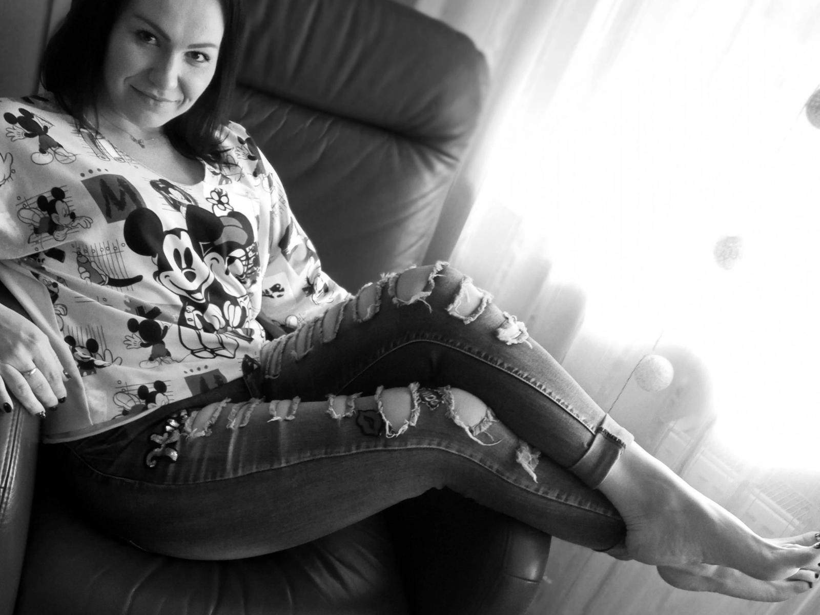 JennyKiss-hot with bare feet, torn jeans, and a Mickey Mouse shirt, sitting in a leather chair, smiling at the camera, sunlight filtering from the right.