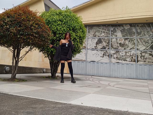 EmmaaRodriguez stands in front of a building, wearing a black strapless top, black shorts, black thigh-highs, and black boots, with a bush to her left.