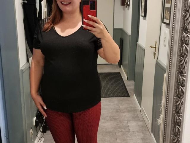 SiegruneValkyra-hot è in un selfie allo specchio, indossa una maglietta nera e pantaloni rossi con una texture particolare, mostra il seno ed è sorridente con il rossetto rosso. La sua mano tiene un telefono rosso..