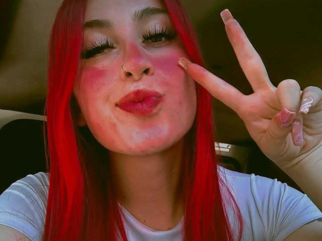 SammyViolet with red hair, nose ring, and pink blush, making a peace sign, puckering her lips, wearing a white shirt, and sporting long, fake nails.