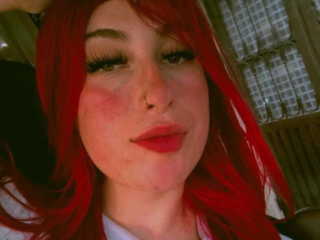 SammyViolet ha i capelli rosso acceso, un piercing al naso e ciglia folte. Sta guardando la fotocamera con le guance leggermente arrossate e rossetto rosso. Il suo seno non è visibile..