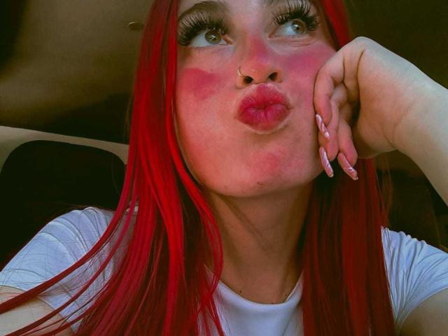 SammyViolet with bright red hair, pouty pink lips, nose ring, and long nails, blowing kiss, wearing white shirt, looking up, in car, her face is flushed.