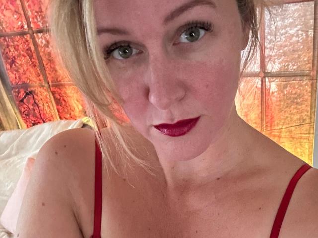 MarleneBloem, con labios rojos, ojos verdes y cabello rubio, vestida con un top rojo de tirantes finos, mirando directamente a la cámara con una ligera sonrisa. Sus senos son parcialmente visibles..