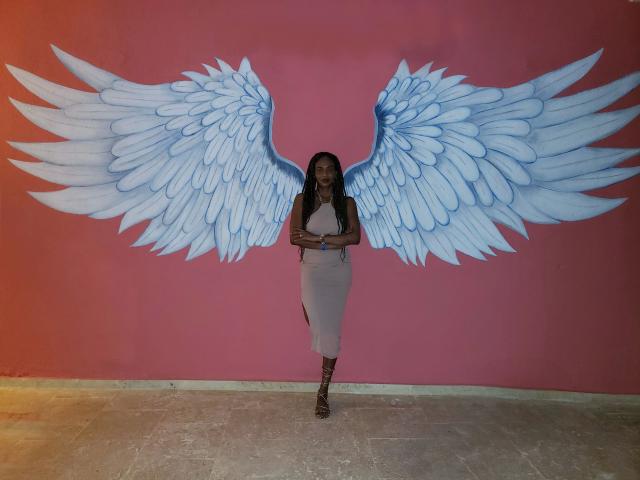 Schoko-Bebe with large blue wings painted on a pink wall, wearing a light pink dress and red lace-up boots, standing on a gray tiled floor.