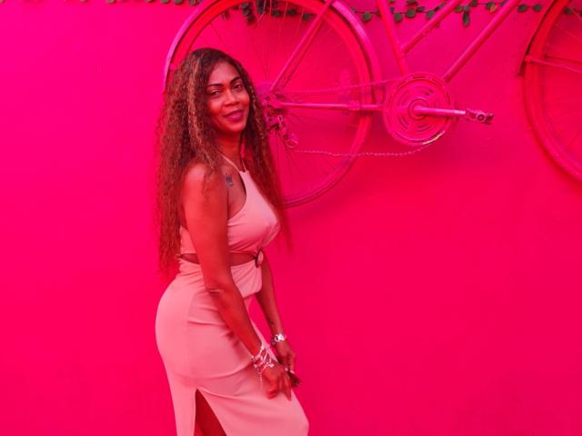Schoko-Bebe with long curly brown hair, wearing a pink dress, standing, smiling, showing her ass with a side slit, against a pink wall with bicycle wheels.