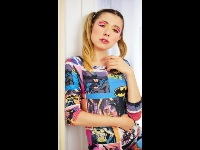 FlexyHeidi-hot with pigtails, pink eye makeup, and colorful Batman-themed dress, standing against white wall, hand near neck, looking slightly annoyed, with a cute yet fierce expression.