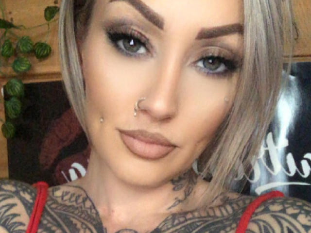 MaitresseLexia has blonde hair, blue eyes, heavy black eyeliner, tattoos on her neck and chest, nose piercings, and a nose ring, wearing a red top.