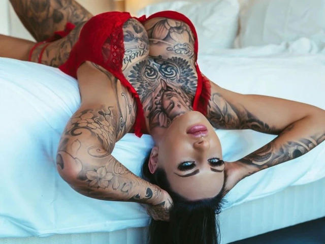 MeganSteel-hot, tattooed from neck to wrists, lying on white bed, red lingerie, arms above head, staring up, large boobs, full lips, dark hair, white pillows, sexy.