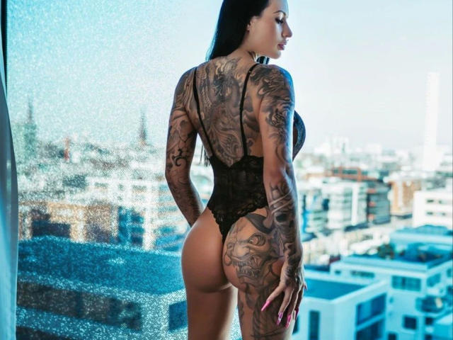 MeganSteel-hot with tattoos, black lace lingerie, and a butt, standing by a window, cityscape outside, looking back, long black hair, pink nails, butt tattoo visible.