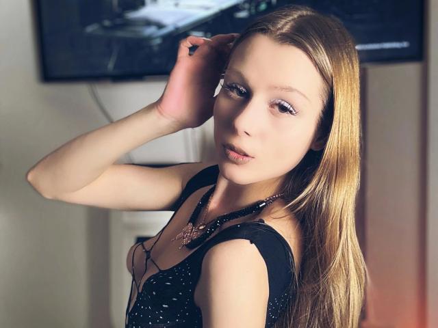 HaneyNanny-hot with long blonde hair, light skin, and pink eyeliner, wearing a black patterned top, looking over her shoulder, hand in her hair, subtle smile, and a necklace.