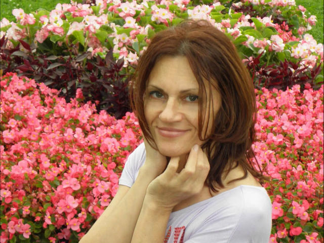 AnnaBronx-hot with red hair, smiling, hands near chin, wearing white shirt, pink flowers in foreground, green grass in background, looking cute, soft light, casual pose, slight smile.