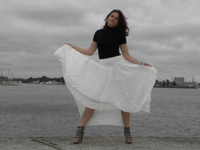 AnnaBronx-hot standing with a flirty smile, black t-shirt, white skirt, and brown strappy sandals, holding skirt out, overcast day, grey sky, waterfront background, short wavy dark hair.