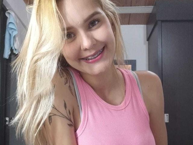 LousaI, rubia, con un piercing en la nariz y un adorno dental, sonriendo con una camiseta rosa, mostrando un tatuaje en el hombro derecho. Tiene pechos pequeños y un trasero lindo..