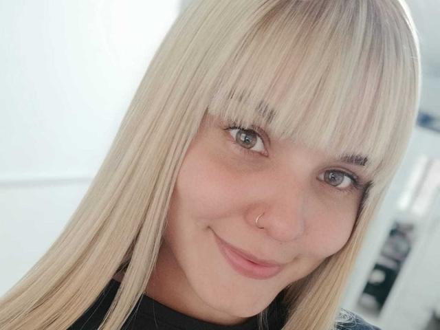 LousaI tiene el cabello rubio y lacio con flequillo, un piercing en la nariz y una sonrisa sutil. Está mirando a la cámara con ojos verde claro y unos labios ligeramente rosados..