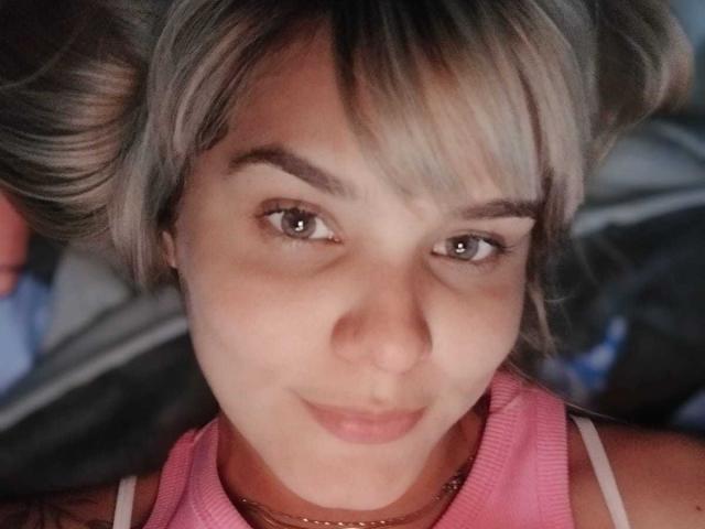 LousaI, avec des cheveux blonds et gris, des yeux marrons, un débardeur rose, souriant légèrement, allongée, éclairage doux, maquillage discret, expression douce, gros plan, détendue, mignonne, attirante..