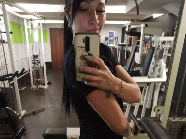 GeiileDeena-hot che si scatta un selfie in palestra, indossa una maglietta nera, ha i capelli lunghi neri raccolti in una coda di cavallo, ha le unghie verdi, tiene in mano un telefono con un design carino, e guarda oltre la spalla..