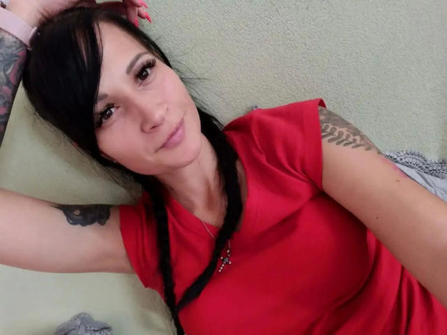 geileDeena - eine attraktive Frau mit schwarzen Zöpfen, rotem Hemd, Tattoos an den Armen, einem rosa Armband, die liegt, mit leichtem Lächeln, blickt in die Kamera, hat rosa Nägel, mittelgroße Brüste und helle Haut..