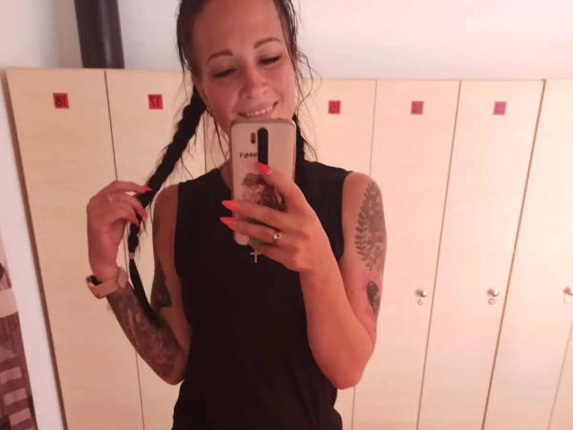 GeileDeena-atractiva con cabello trenzado, top negro sin mangas, brazos tatuados, uñas rojas, sonriendo, sosteniendo un teléfono, selfie en el espejo, mostrando los senos, sosteniendo el trenzado, anillo en el dedo, en el vestuario..
