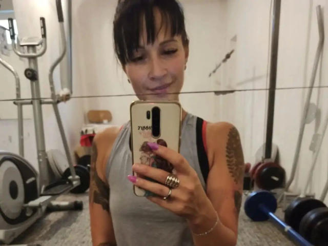 geileDeena-sexy, con tatuaggi, canotta grigia, unghie rosa, si scatta un selfie in palestra, sorridente, tiene il telefono, sedere visibile, spallina del reggiseno rosa, anello al dito, capelli castani raccolti in uno chignon..