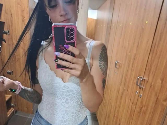 geiileDeena, capelli neri raccolti in una coda di cavallo, top di pizzo bianco, seno visibile, tatuaggi sul braccio destro, tiene in mano un telefono rosa, in piedi in una stanza di legno..