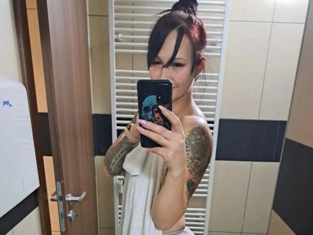 Autoportrait dans un miroir de geileDeena-hot, avec des cheveux roux et noirs, des tatouages sur le bras droit, tenant un téléphone qui cache ses petites seins avec une serviette blanche, debout dans une salle de bain..