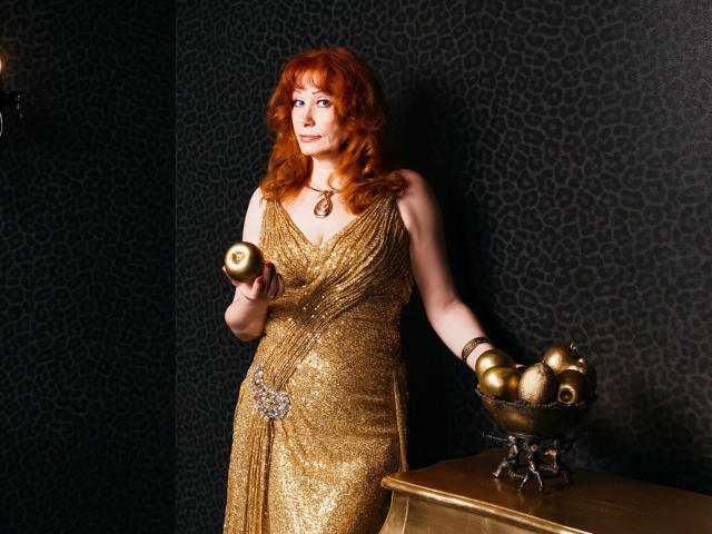 MomAimy-hot with red hair, wearing a shimmering gold dress, holding a golden apple, standing next to a brass bowl with more apples, in a dark room.