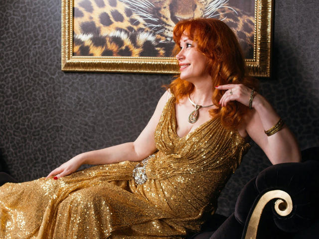 MomAimy-hot with red hair, gold sequin dress, and necklace, sitting on black velvet couch, smiling, pointing at leopard painting, wearing bracelet, looking to the side.