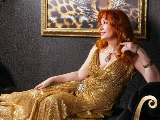 MomAimy-hot with red hair, wearing a glittery gold dress, sits on a black chair, smiling, hand near face, gold necklace, leopard print painting in background.